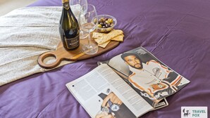 A bed with a purple blanket, a wooden tray with a bottle of Nozeco wine, glasses, olives, crackers, and a magazine featuring a man in a white shirt.