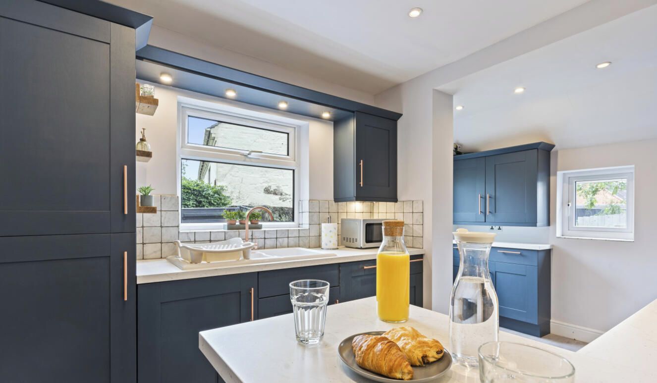 Et moderne køkken med mørke skabe, en hvid bordplade, et vindue og et morgenmåltidsbord med croissanter og juice.