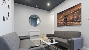 Een moderne woonkamer met grijze banken, een bijzettafeltje met ontbijtartikelen, een ronde spiegel en een stadslandschap schilderij.