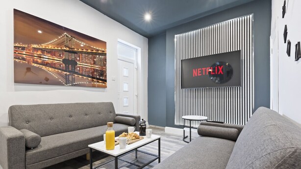 Een moderne woonkamer met een grijze bank, een salontafel met ontbijtartikelen, een groot televisiescherm waarop het Netflix-logo te zien is, en een stadslandschap schilderij aan de muur.