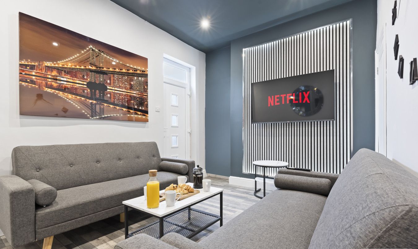 A modern living room with a grey sofa, a coffee table with breakfast items, a large TV screen displaying the Netflix logo, and a cityscape painting on the wall.