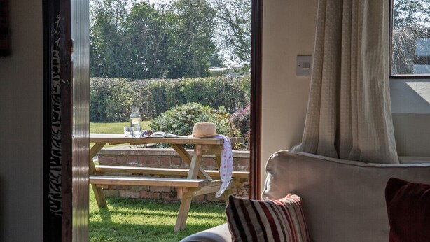 Een knus vertrek met uitzicht op een tuin, een houten tafeltje met een hoed en een boek, en een gestreept kussen op een bank.