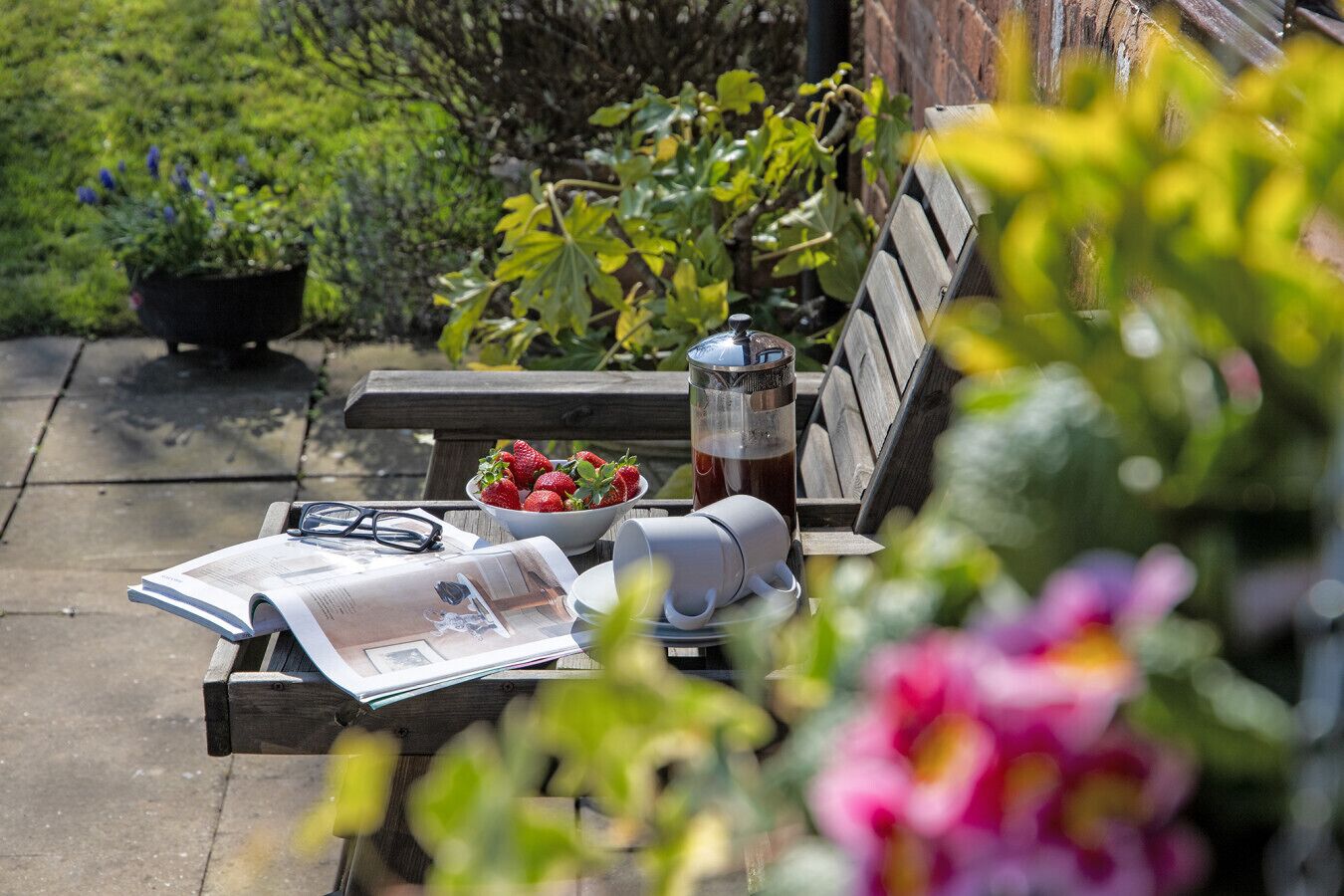A wooden bench with a newspaper, a bowl of strawberries, a thermos, and a glass of iced tea.