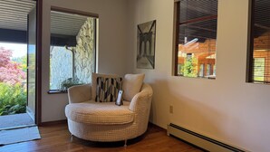 Een hoekappartement met een rond, gestructureerd fauteuil, een groot raam met jaloezieën, een aan de muur hangend schilderij en een houten vloer.