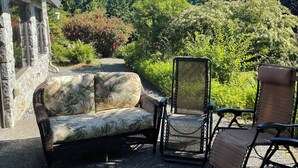 Een terras met een bank en stoelen, een stenen gebouw en een tuin met diverse planten en bomen.