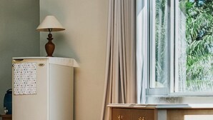 A compact room with a small white refrigerator, a lamp on top, a wooden table with drawers, and a window with curtains. - Nirmala Beach Hotel and Resort Biak (Biak)