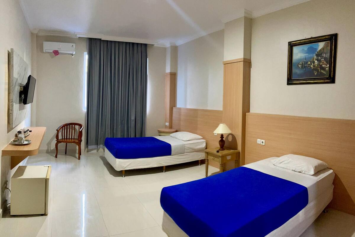 A hotel room with two beds, a desk, a chair, a lamp, and a painting on the wall.