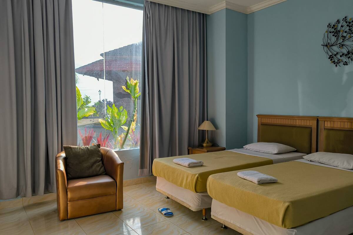 A hotel room with two beds, a chair, a lamp, and a view of the outdoors.