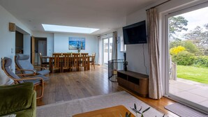 A spacious living area with a dining table, chairs, a sofa, a TV, and a large window with a view of greenery.