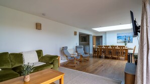A modern living room with a green sofa, wooden coffee table, and two blue armchairs. A dining area with a wooden table and chairs is visible in the background.