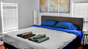Cama bem arrumada com roupa de cama azul e branca, um quadro com mapa mundi acima e uma luminária de cabeceira.