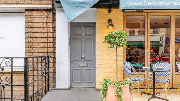 Une devanture de magasin avec une banderole sur le site web, une porte, un petit arbre et des sièges d’extérieur avec des chaises à rayures.