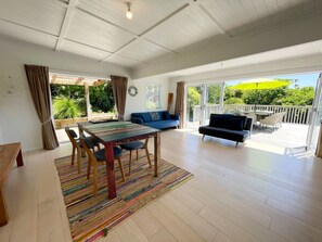 A spacious living area with a wooden table, blue sofa, and a view of the outdoors through large sliding doors. - Oneroa Village Oasis - Central Near Beaches (Waiheke Island)