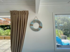 Ein Zimmer mit beigen Vorhängen, einer Wanddekoration im Strandstil und Blick auf einen Pool durch ein Fenster mit Jalousien.
