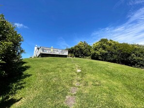 A grassy slope leading to a white building with a balcony, surrounded by trees.
