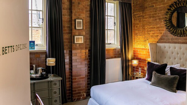 A hotel room with a brick wall, a bed with white linens, a headboard, a mirror, a dresser, a lamp, and a window with curtains. - Betts Private room at Bicester Heritage (Bicester)