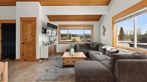 A modern living room with a wooden ceiling, a large window with a mountain view, a gray sofa, and a wooden coffee table.