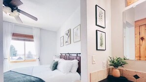A bedroom with a bed, a ceiling fan, a window with curtains, and framed artwork on the wall.
