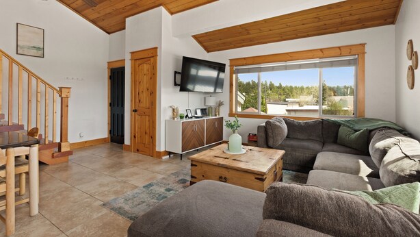 A spacious living area with a wooden ceiling, a large window offering a view of the outdoors, a television mounted on the wall, and a cozy seating arrangement.