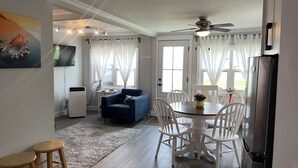 Un coin repas avec une table ronde, des chaises blanches, un ventilateur de plafond et un réfrigérateur.