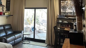 A modern living room with a black leather sofa, a wooden dining table, and a large window with curtains.