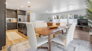 A modern kitchen with dark cabinets, a stainless steel refrigerator, and a dining area with a wooden table and white chairs. - Urban Oasis with Cedar Hot Tub (Seattle)