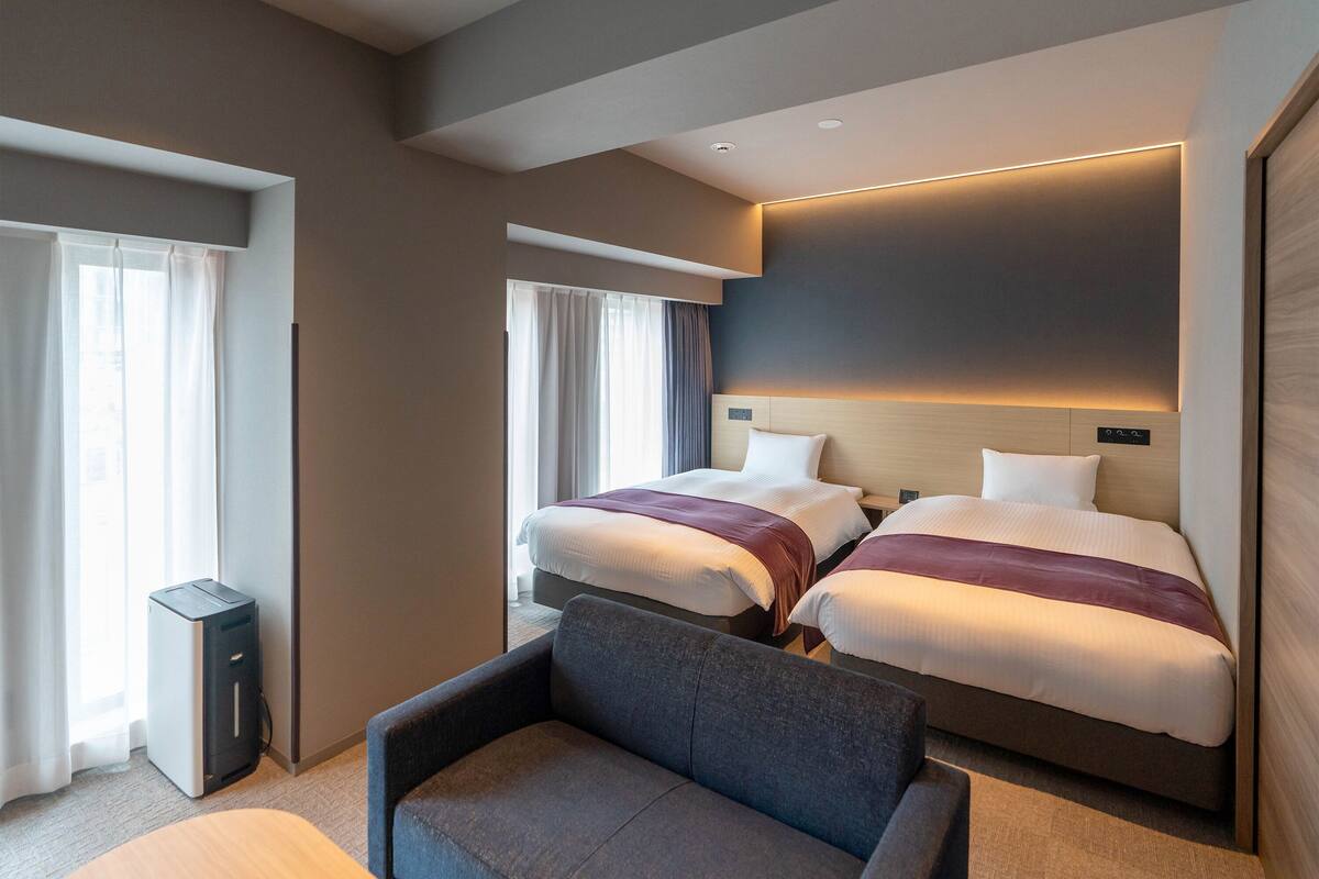 Hotellihuone, jossa on kaksi sänkyä, sohva, työpöytä ja suuri ikkuna.