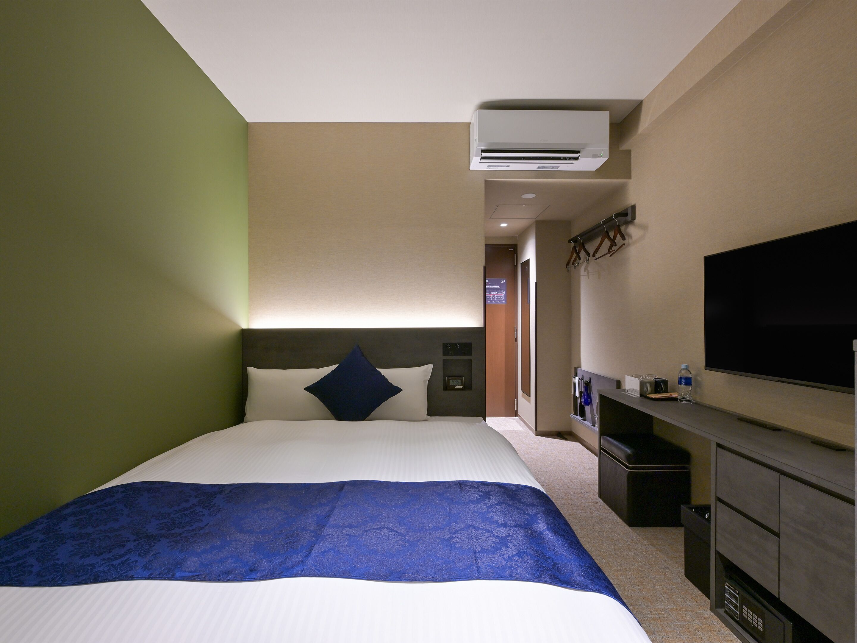A hotel room with a bed, a TV, an air conditioning unit, a desk with a chair, and a wardrobe.