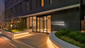 A modern hotel entrance with a glass door, a well-lit pathway, and a landscaped garden area. - HOTEL R9 Premium Ichikawaekimae (Ichikawa)