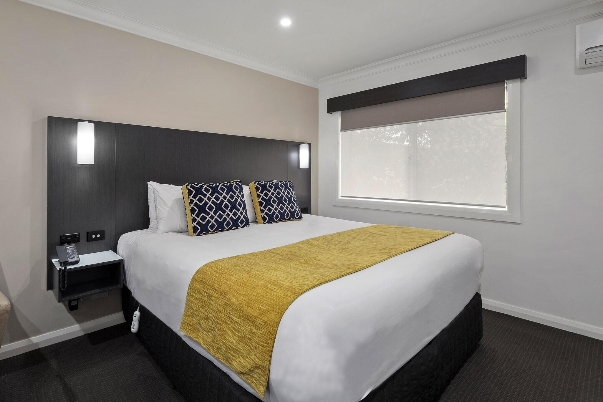 Habitación de hotel con una cama grande, una mesita de noche con teléfono, una ventana con persianas y una lámpara de pared.