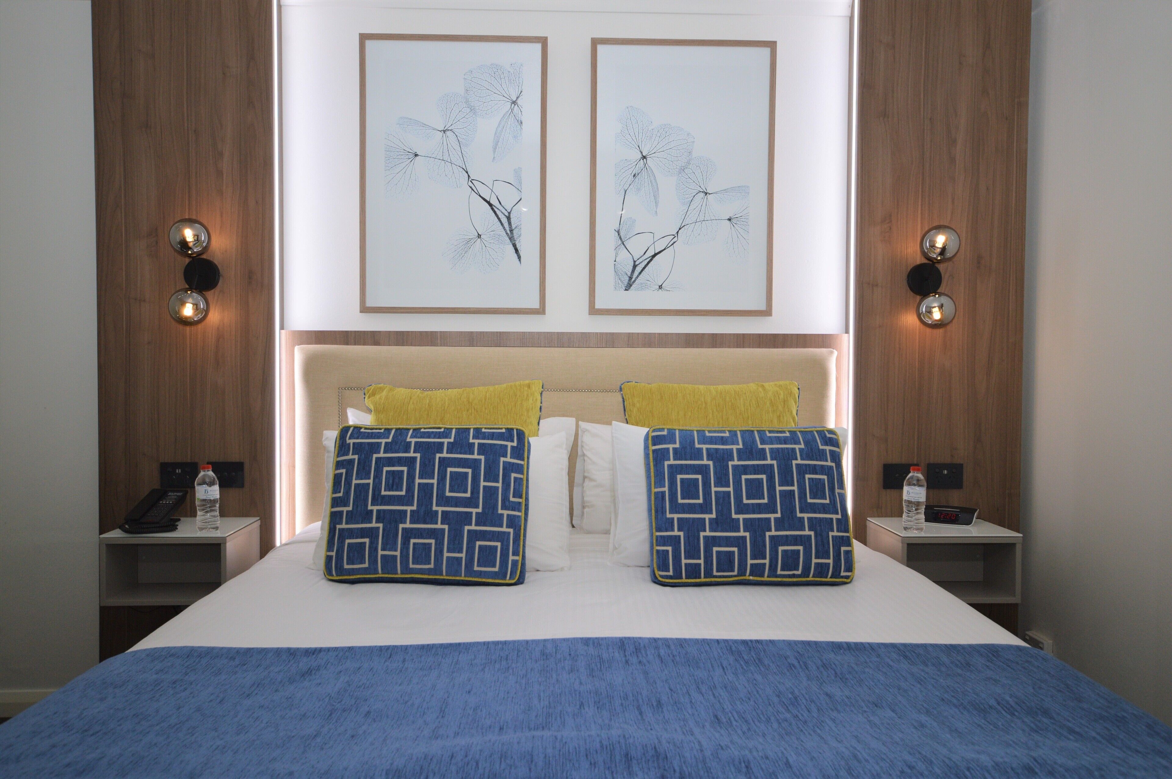 A hotel room with a bed, two wall-mounted light fixtures, a bedside table with a phone and a bottle, and two framed wall art pieces.
