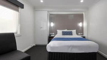 Una habitación de hotel con cama, una silla, un armario y una mesita de noche con lámpara.