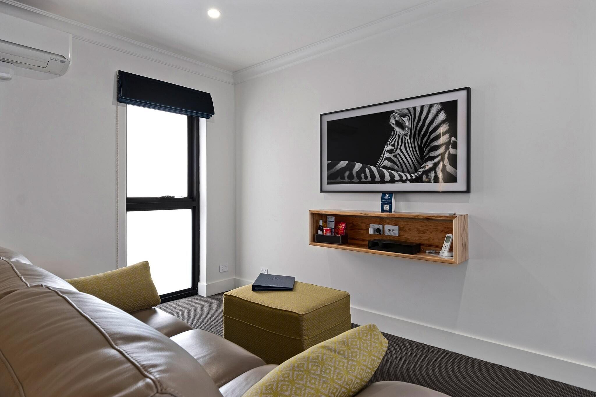 A modern living room with a leather sofa, a small ottoman, a framed zebra artwork, a wooden shelf with items, and a window with a black roller blind.