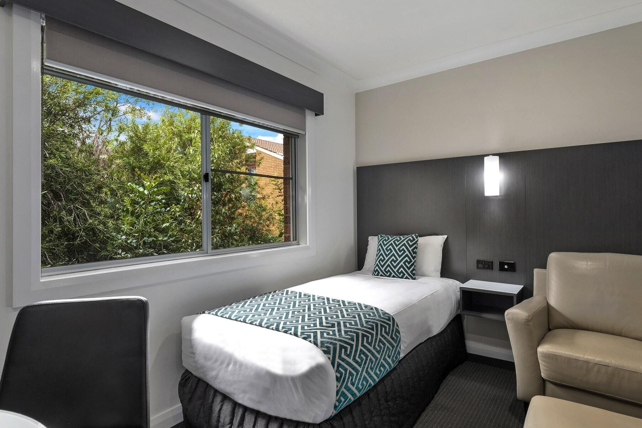 A modern hotel room with a single bed, a chair, a nightstand, and a window with a view of trees.