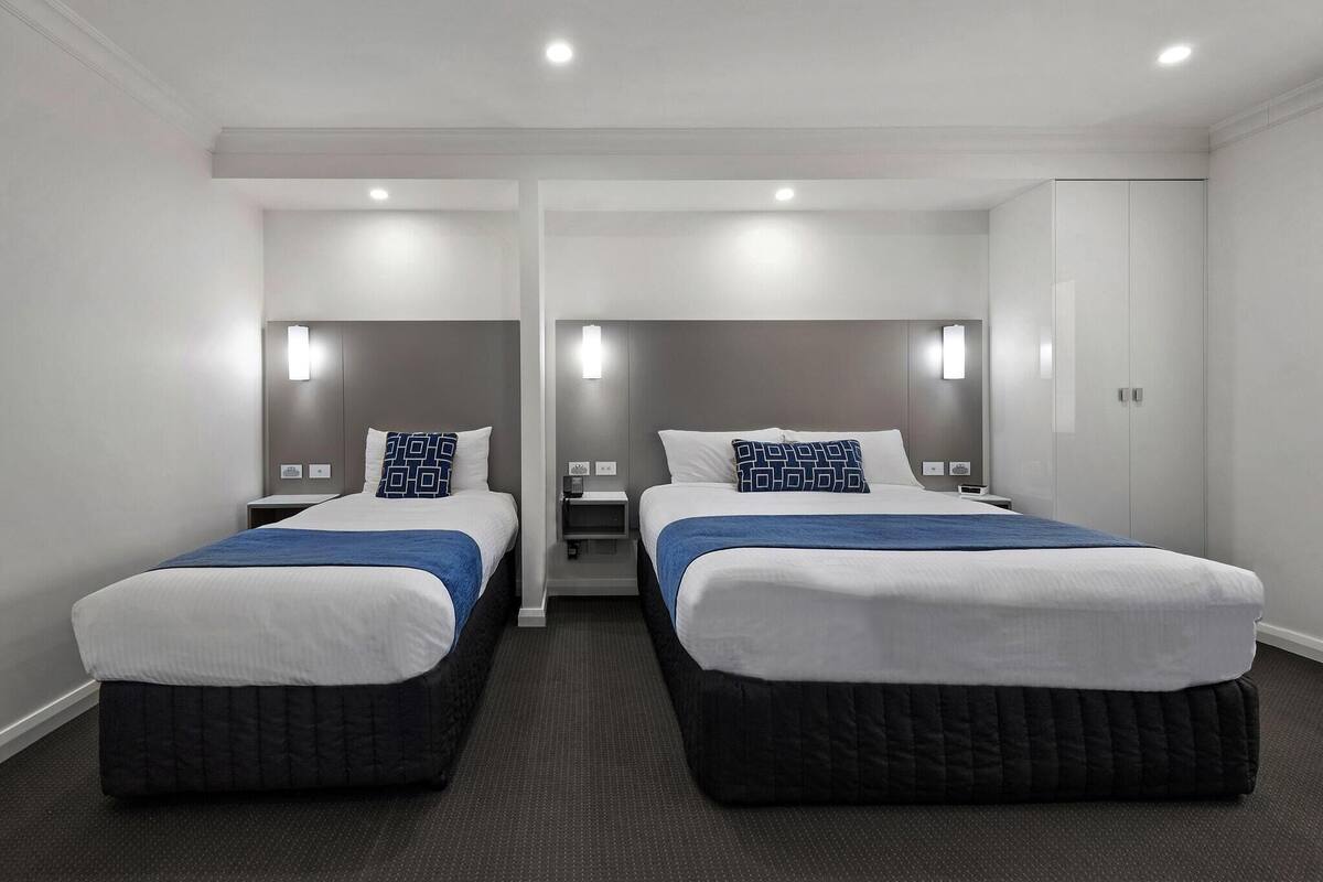 A hotel room with two beds, each with a white and blue bedspread, a dark headboard, and a nightstand with a lamp.