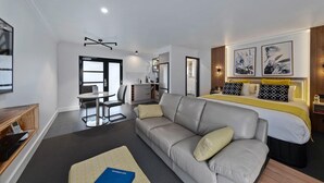 Une chambre d’hôtel moderne avec un canapé gris, un lit avec une literie jaune et noire, un bureau avec une chaise et une petite kitchenette.