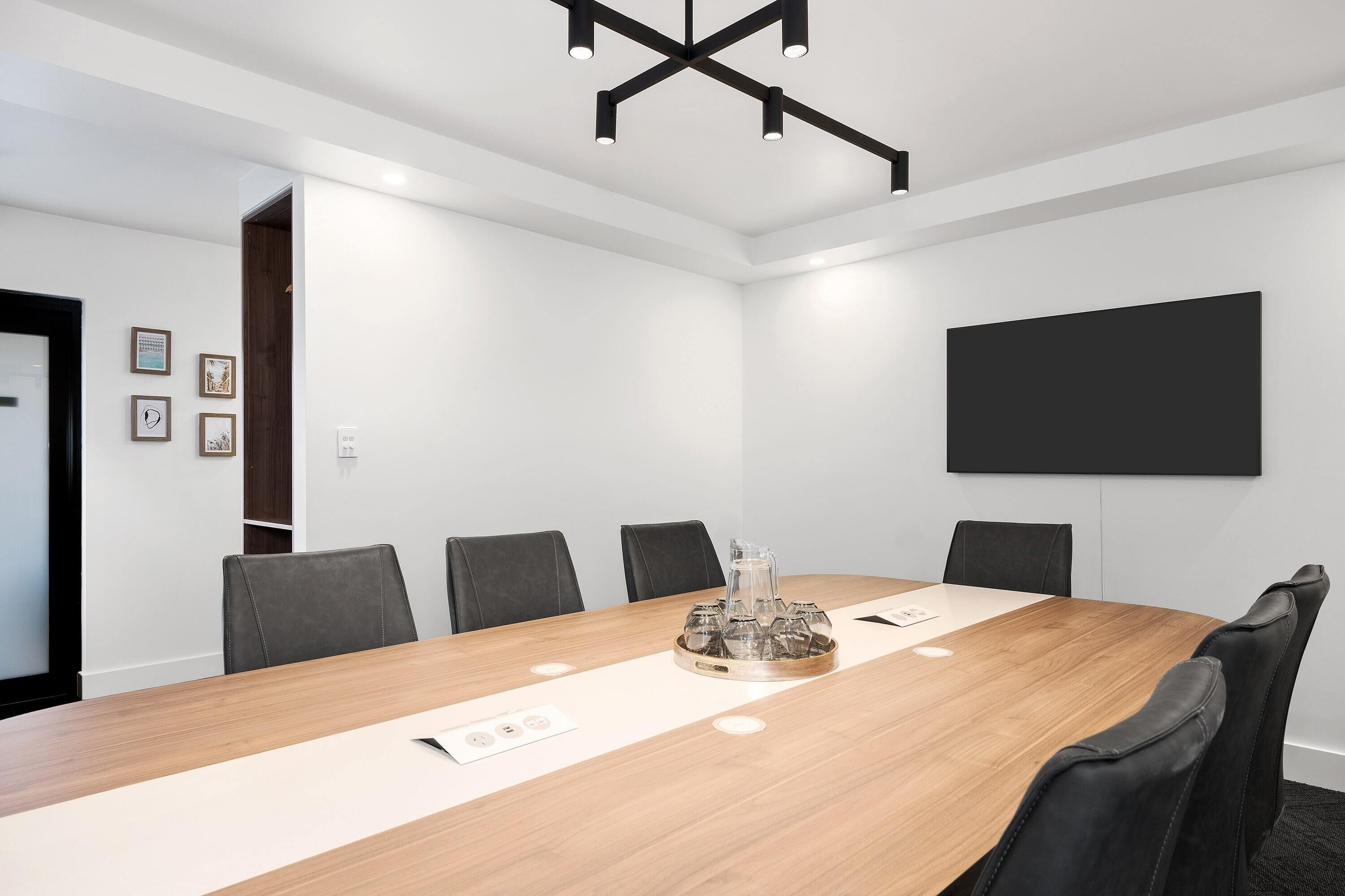 A conference room with a long wooden table, black chairs, a projector screen, and a modern ceiling light fixture.