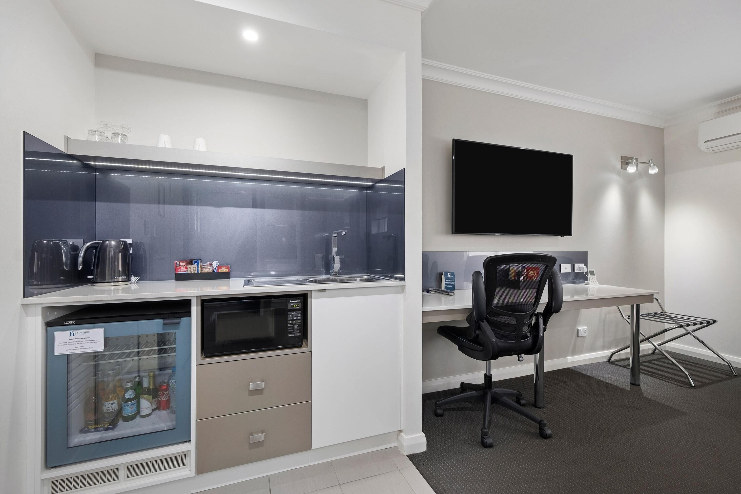 A modern kitchen with a built-in refrigerator, microwave, and kettle. There is a flat-screen TV mounted on the wall and a black office chair.