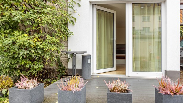 A modern outdoor area with a sliding glass door leading to a room, surrounded by greenery and planters with flowering plants.