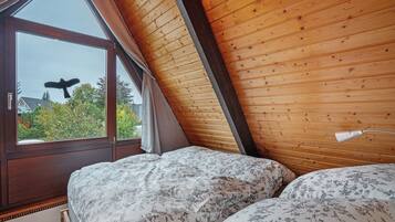 Ein Schlafzimmer mit schrägen Wänden, Holzboden und einem großen Fenster mit Vorhängen. Ein Bett ist mit floralem Bettbezug ausgestattet.