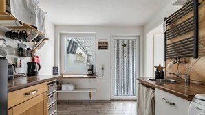 A modern kitchen with wooden cabinets, a built-in oven, a coffee machine, and a window with a curtain. - Holiday Home 'Nilsson' with Private Terrace, Private Garden and Wi-Fi (Heide)