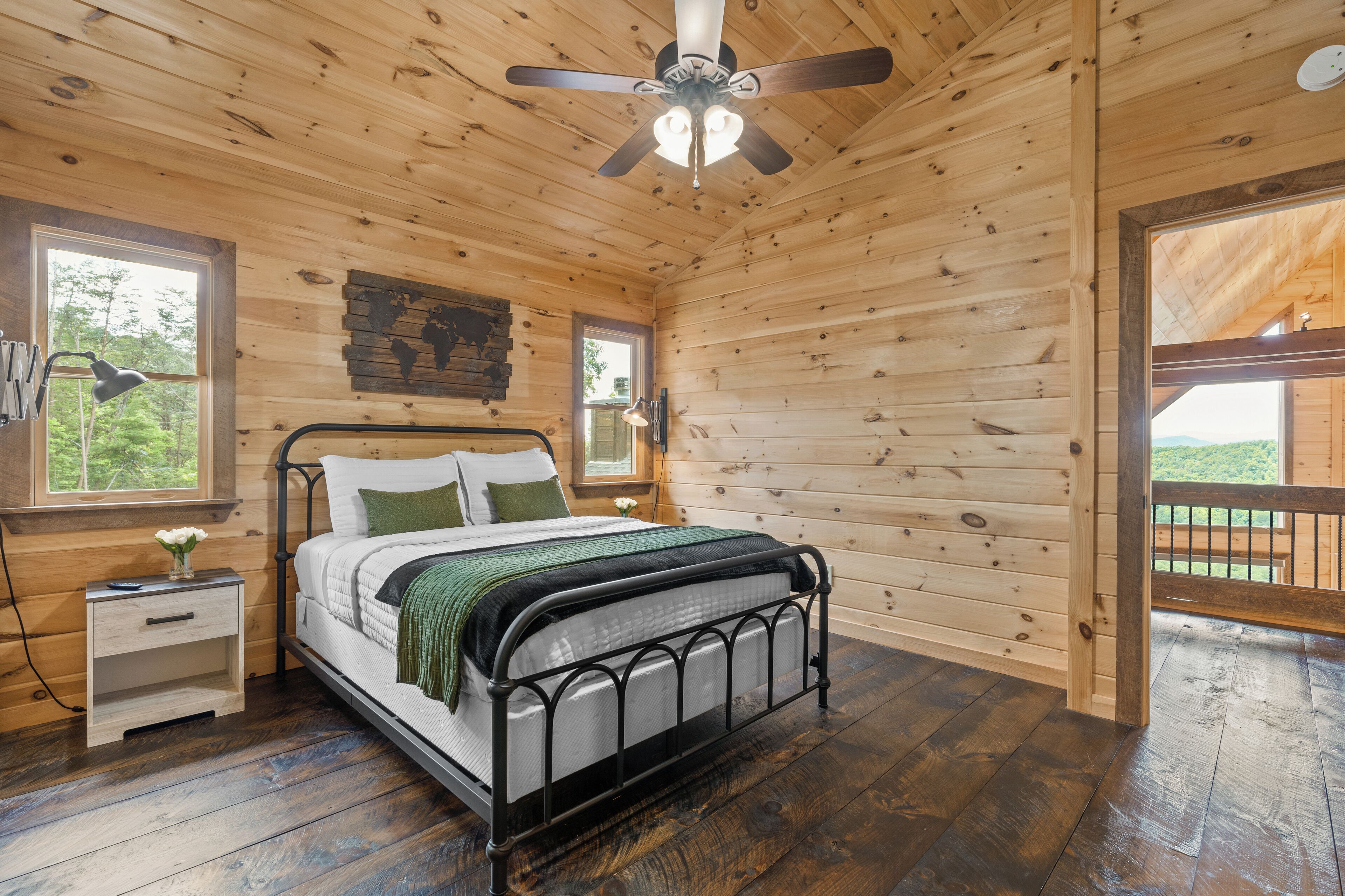 A wooden cabin bedroom with a metal bed frame, a nightstand, a desk lamp, and a ceiling fan.