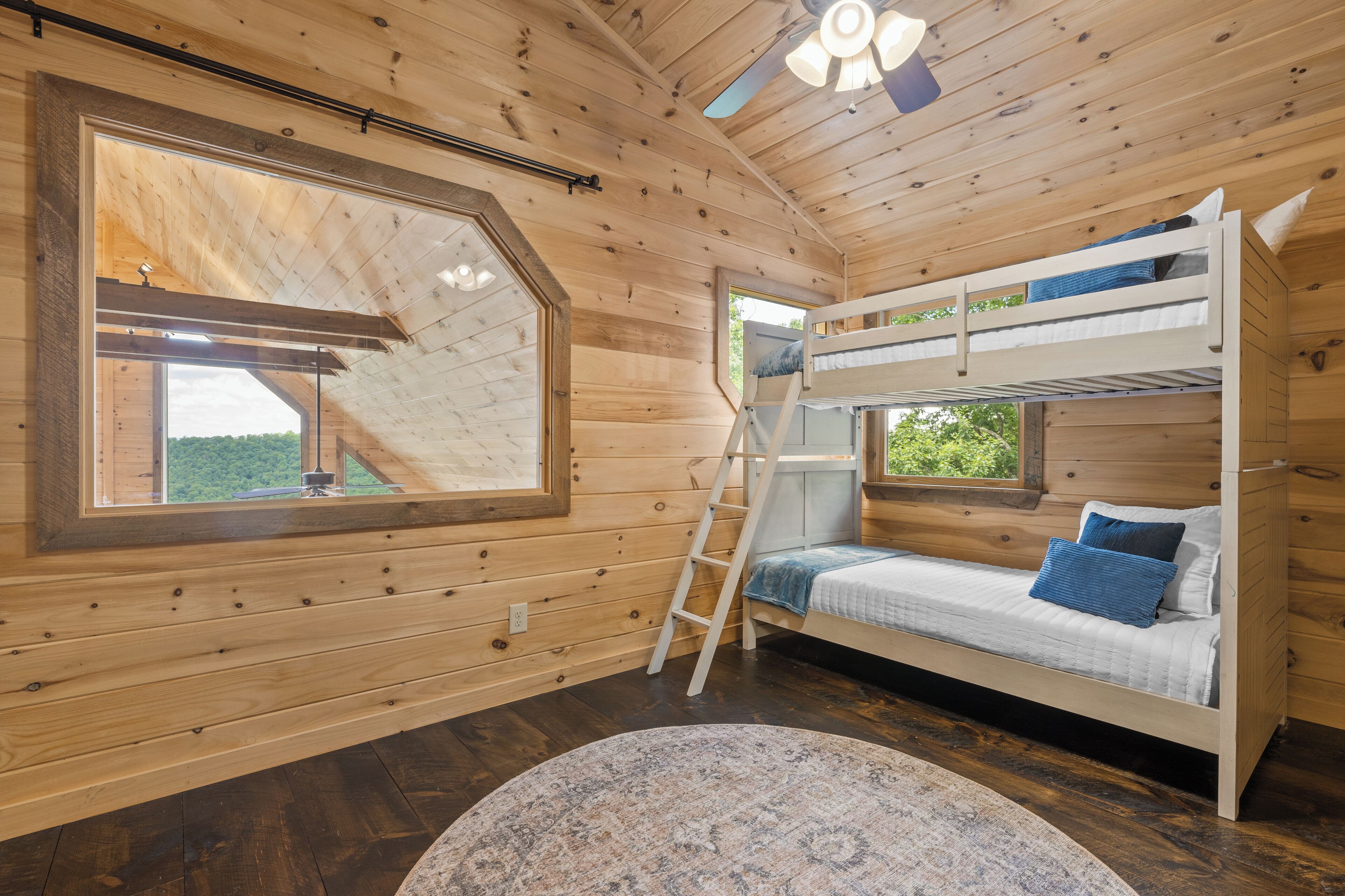 A wooden cabin room with a bunk bed, a ladder, a large window, and a ceiling fan.