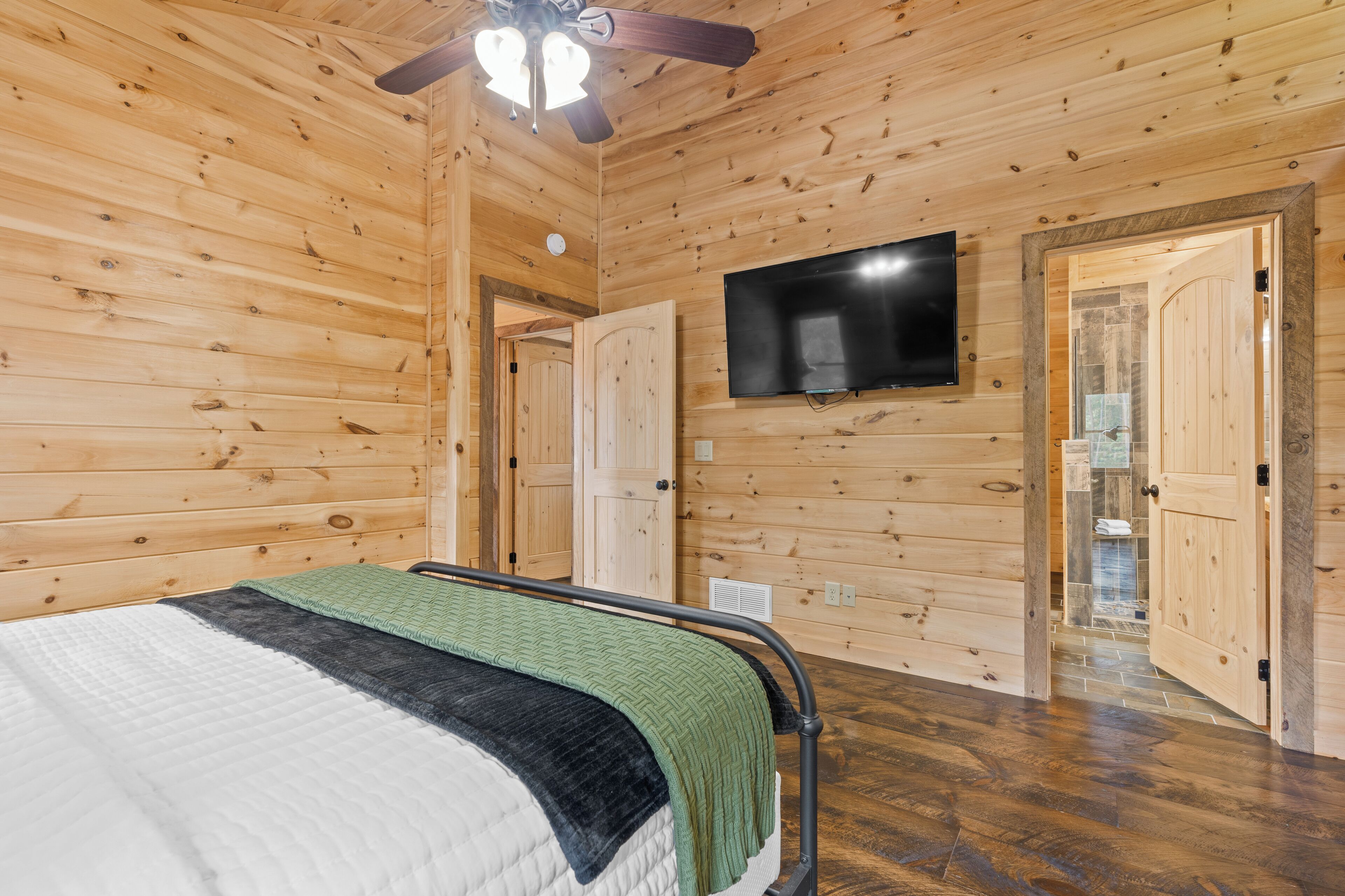 A wooden-paneled room with a bed, a ceiling fan, a flat-screen TV, and a bathroom door.