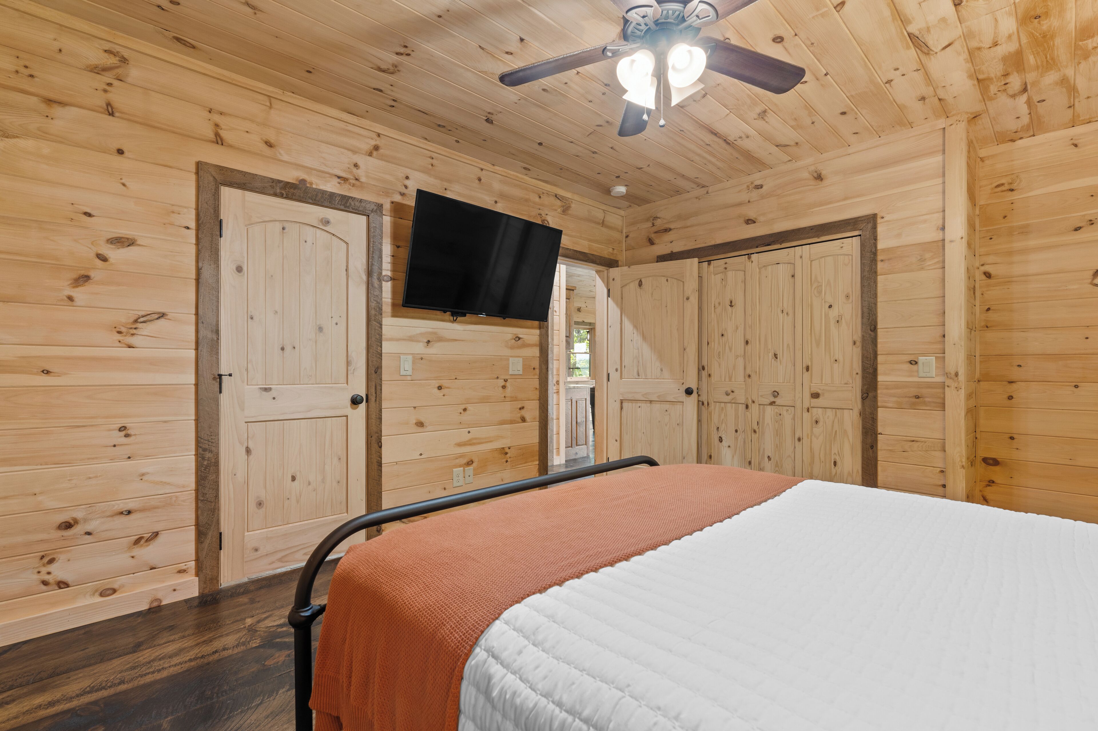 A wooden-paneled room with a bed, a ceiling fan, and a television mounted on the wall.