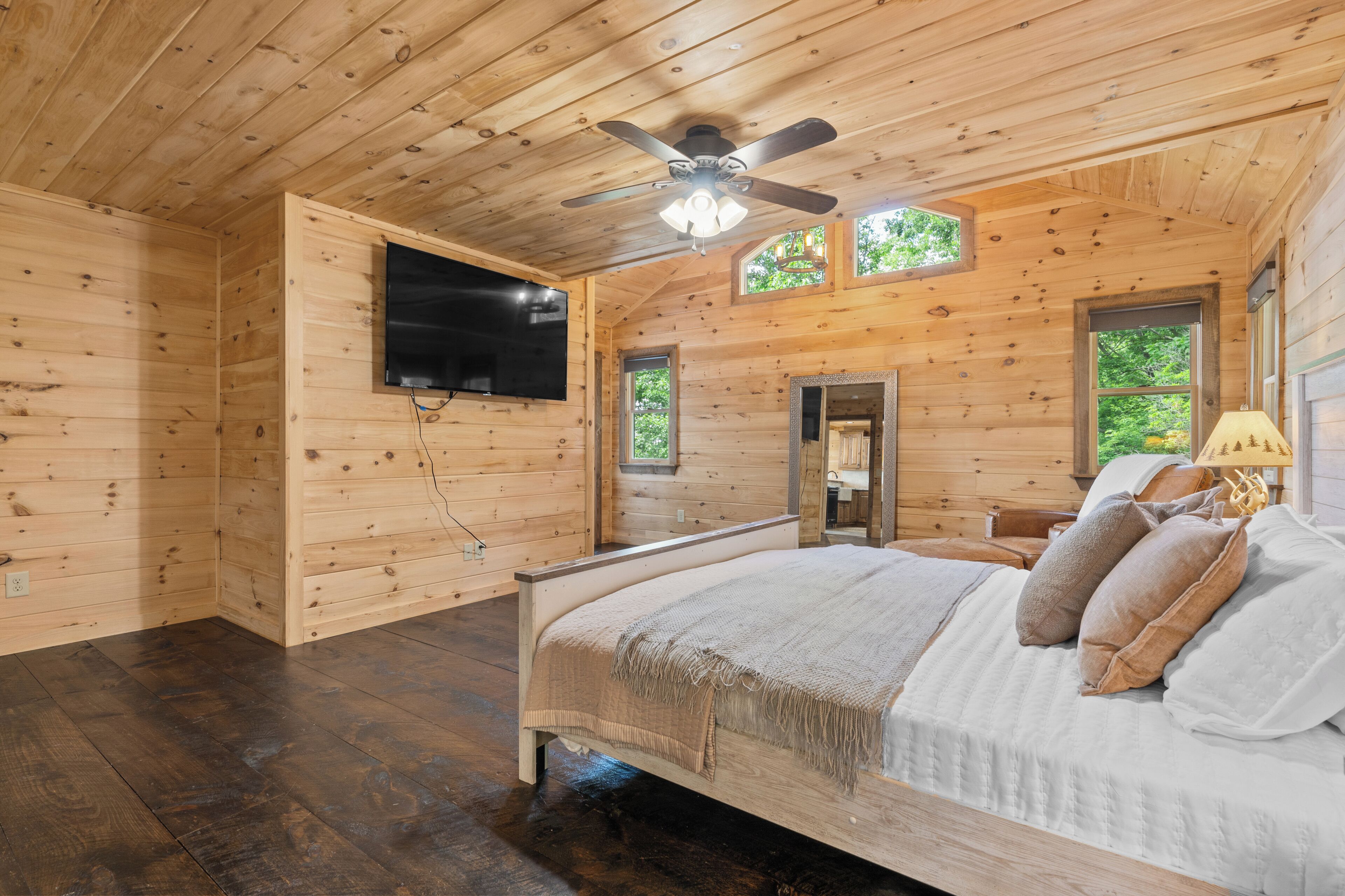 A wooden cabin bedroom with a bed, ceiling fan, TV, and windows.