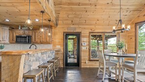 A rustic wooden interior with a stone counter, wooden stools, a dining table with chairs, and a black-framed door leading outside. - Serenity View (Blue Ridge)
