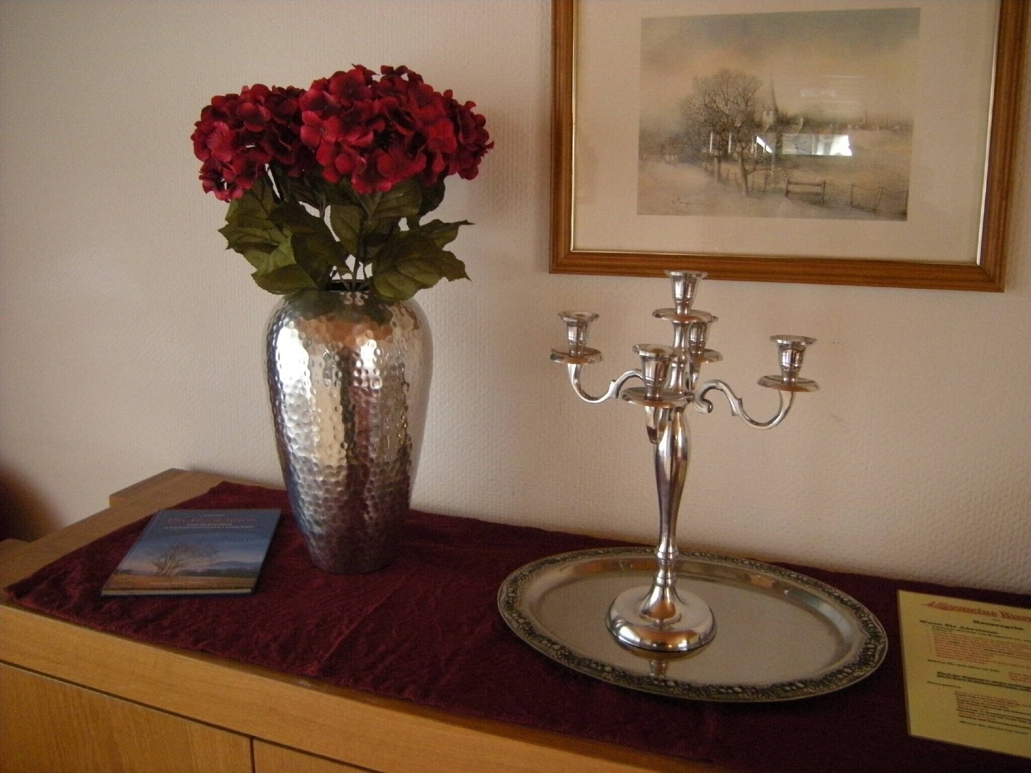 A wooden desk with a vase of red flowers, a framed picture, and a candelabra.