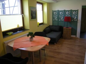 Une pièce comprenant une table, un canapé, une fenêtre avec des stores, une plante en pot et un élément de séparation décoratif.