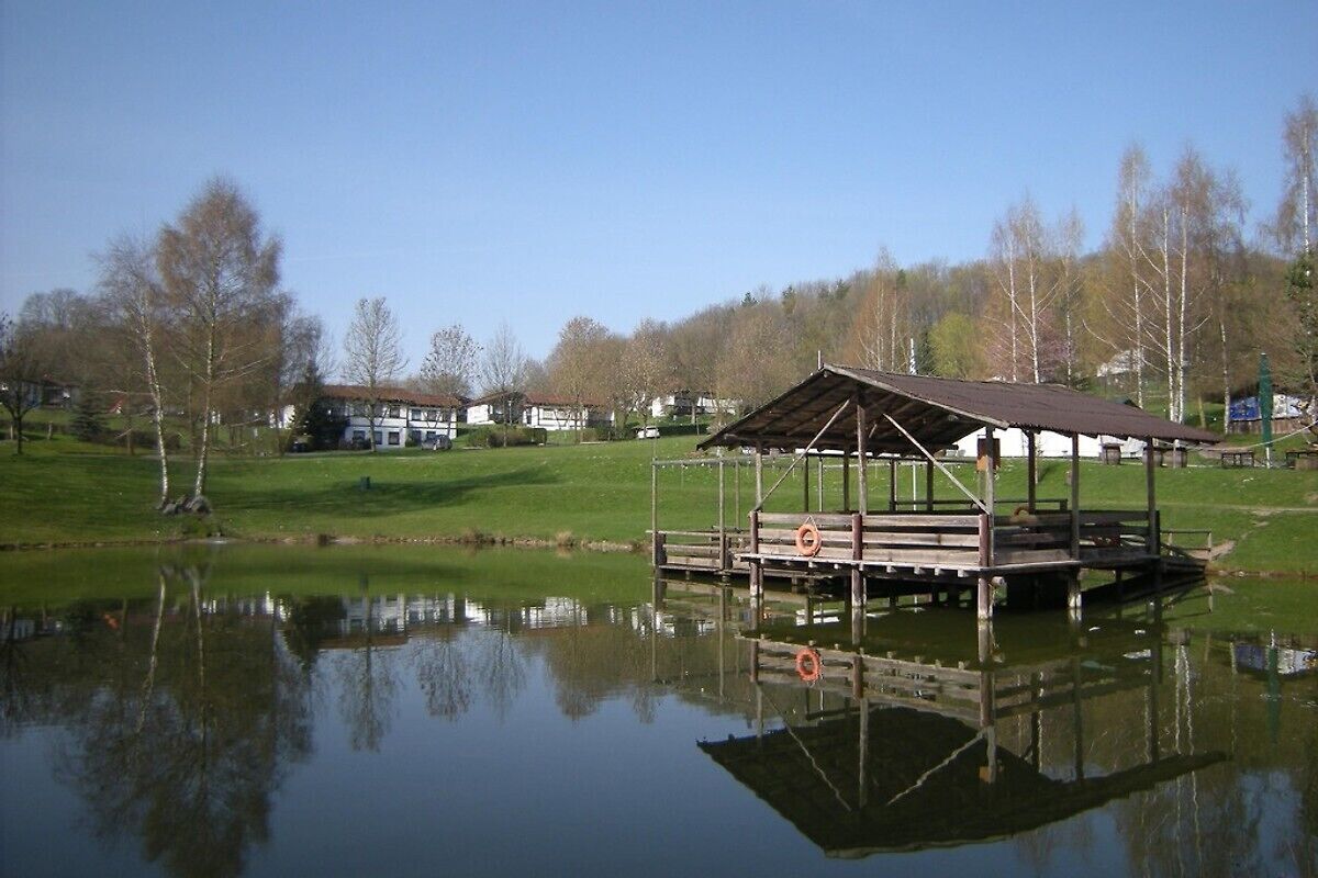 Une jetée en bois surmontée d’un abri, surplombant un lac paisible, entourée d’arbres et avec des maisons en arrière-plan.